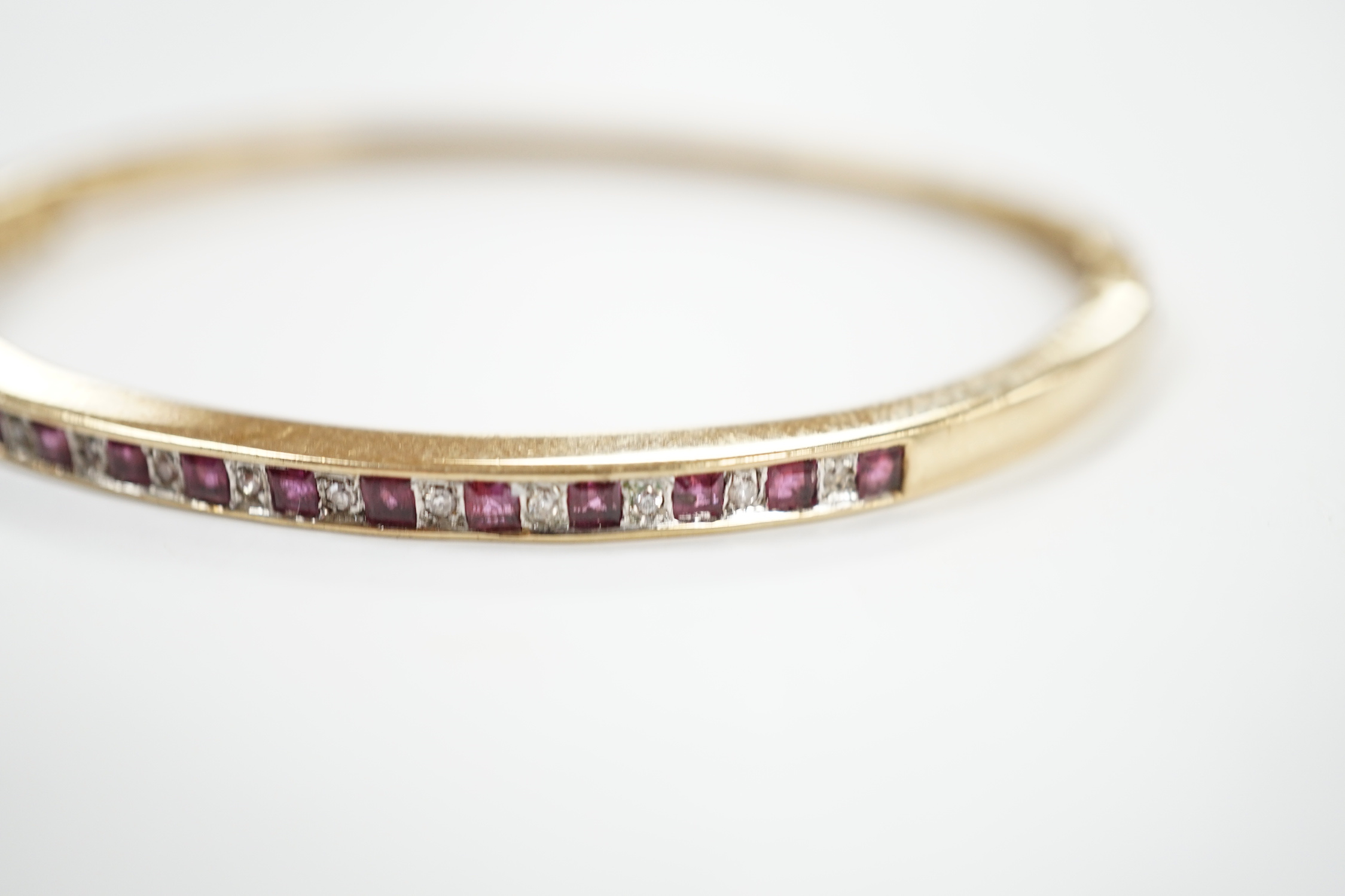 A yellow metal, ruby and diamond chip channel set hinged bangle, interior diameter 57mm, gross weight 10.8 grams.